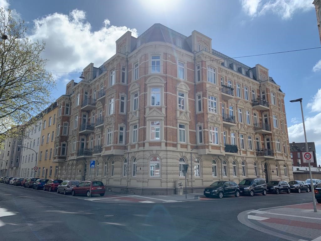 Ein imposantes Eckhaus im Stil der Gründerzeit, frisch renoviert mit sauberer Fassade und restaurierten Stuckelementen. Die Sonne scheint von oben ins Bild, wodurch ein leichter Lichtschein sichtbar ist. Auf der Straße stehen zahlreiche Autos, und ein Verkehrszeichen weist auf eine Tempo-30-Zone hin.